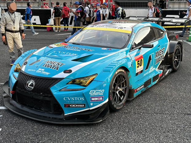 Blauer Lexus RC F GT3 Rennauto auf einer Straße mit einer Gruppe von Menschen, einem Verkehrskegel und Hintergrundbeschilderung.