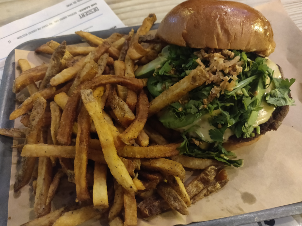 Ein Tablett mit einem Hamburger, der mit Salat, Tomate und Zwiebeln belegt ist, und goldenen, knusprigen Pommes frites, das auf einem Tisch mit einem Blatt Papier daneben steht.