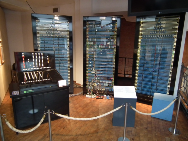 Ein unordentlicher Raum mit verschiedenen elektronischen Geräten, ein Tisch mit Gegenständen, eine bodengestützte Tafel mit Text, Absperrungen aus Seilen auf Pfählen, ein Metallzaun, gerahmte Fotos an der Wand und Deckenleuchten.