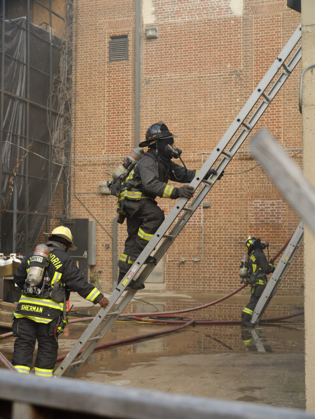 Feuerwehrleute in Helmen und Ausrüstung klettern an einer Leiter an einem Backsteingebäude mit Rohren auf dem Boden und einem Metallstab an der Basis vorbei, während im Hintergrund ein weiteres Gebäude mit Fenstern und einem Netz zu sehen ist.
