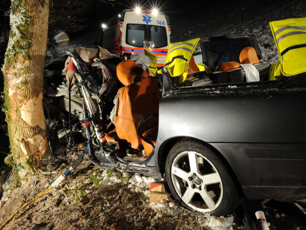 Ein verunglütztes Auto am Rande einer schneebedeckten Straße mit einem im Hintergrund sichtbaren Krankenwagen.