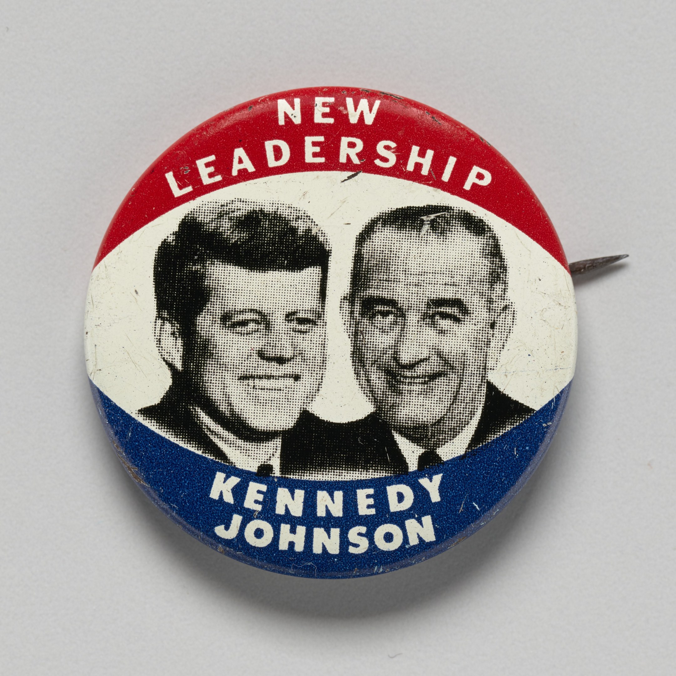 Eine weiße Oberfläche mit einem Ansteckbutton mit zwei Bildern von Kennedy und Johnson und der Aufschrift "Neue Führung" darunter.