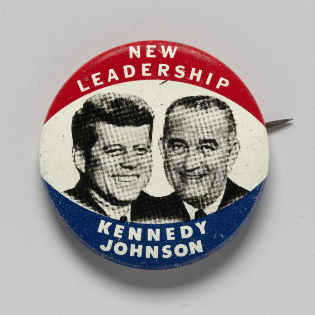 Eine weiße Oberfläche mit einem Ansteckbutton mit zwei Bildern von Kennedy und Johnson und der Aufschrift "Neue Führung" darunter.