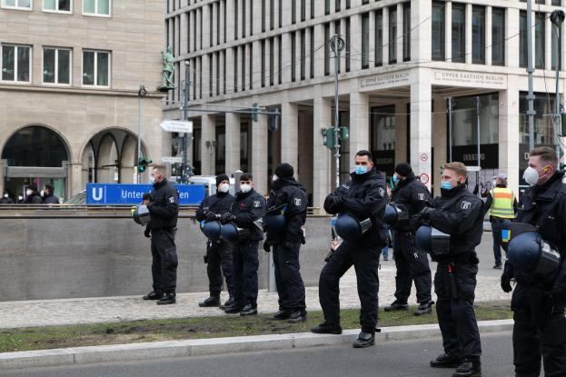 Eine Gruppe von Polizisten in schwarzen Uniformen und Masken steht vor einem Glasfenstergebäude mit Säulen in Berlin, Deutschland, einige halten Helme, mit Laternen, Verkehrsampeln, Texttafeln, einer Statue, anderen Menschen und grasbewachsenem Boden im Hintergrund.