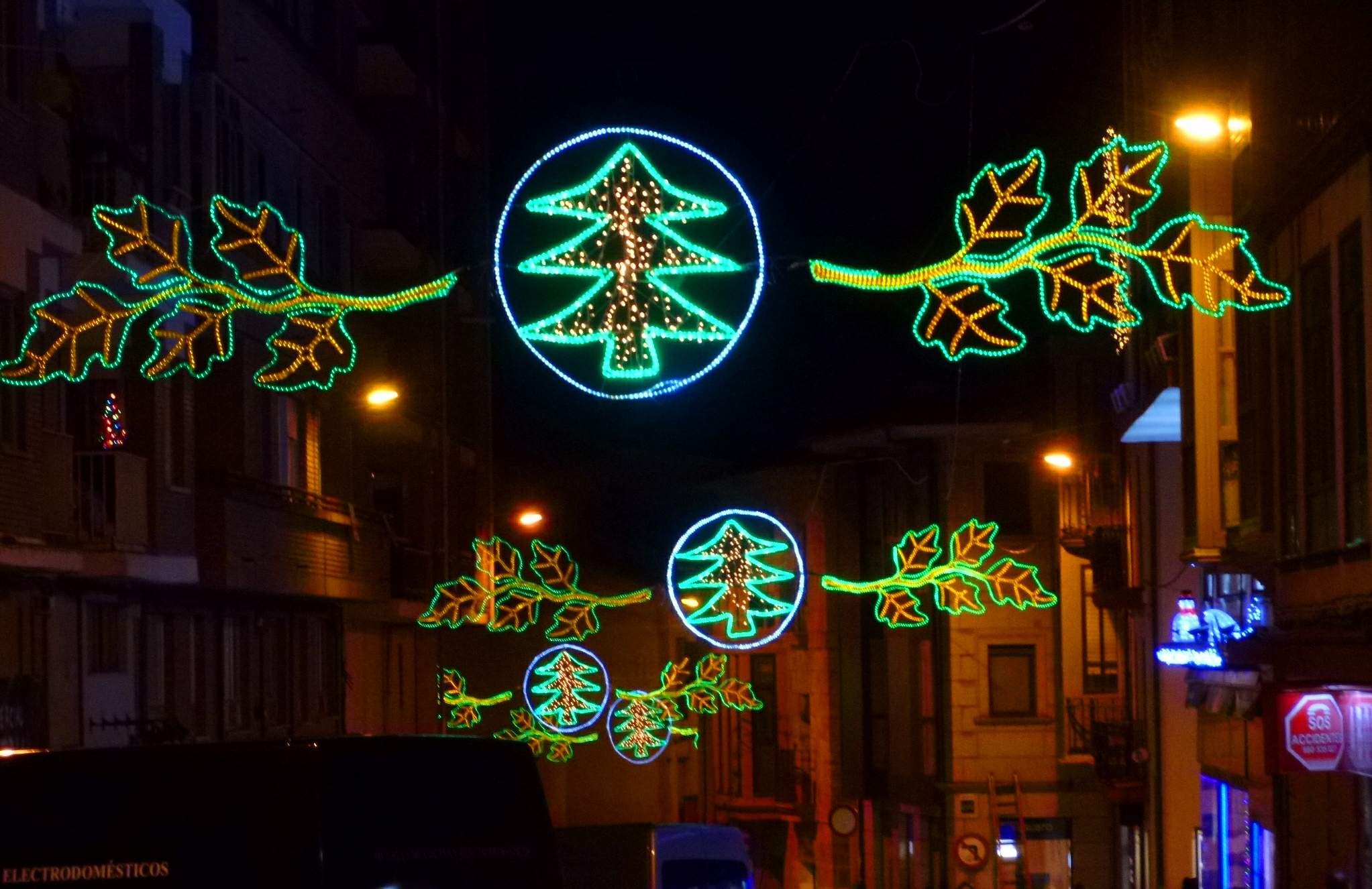 Festliche Weihnachtsbeleuchtung mit dekorativen Blättern schmückt eine von Gebäuden gesäumte Stadtstraße, während Fahrzeuge vorbeifahren.