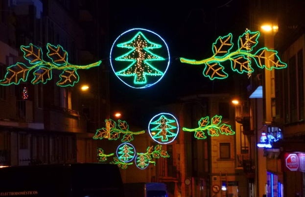 Festliche Weihnachtsbeleuchtung mit dekorativen Blättern schmückt eine von Gebäuden gesäumte Stadtstraße, während Fahrzeuge vorbeifahren.