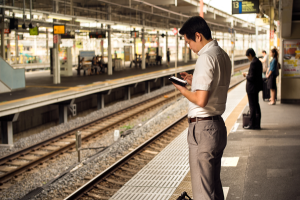 Ein Mann steht auf einem Bahnsteig und schaut auf sein Handy, umgeben von Menschen, mit Bahnschienen im Hintergrund und einem Textschild auf der rechten Seite.