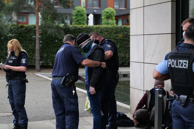 Gruppe von Polizeibeamten vor einem Gebäude stehend, mit einer Frau, die ein Buch und einen Stift hält, umgeben von sitzenden und stehenden Personen, mit Bäumen und Gebäuden im Hintergrund.