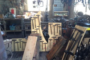 Ein mit Werkzeugen, Geräten und Holzplanken gefüllter Werkstattraum mit einem Stuhl links, Werkzeugen an der Wand und einem Fenster im Hintergrund.