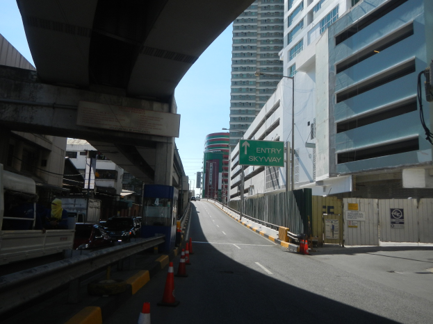 Stadtstraße mit Verkehrskegeln, Fahrzeugen, einer Brücke auf der linken Seite, Gebäuden mit Namenschildern, Laternenpfählen und einem Himmel als Hintergrund.