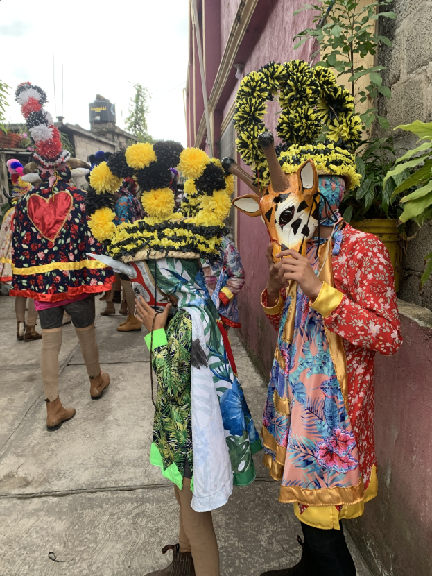Eine Gruppe von Menschen in bunten Kostümen steht zusammen im Freien und hält verschiedene Gegenstände, mit Bäumen, Gebäuden und einem klaren blauen Himmel im Hintergrund.
