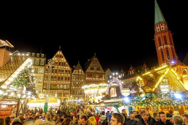 Ein lebendiger Weihnachtsmarkt in Nürnberg, Deutschland, mit Menschen um geschmückte Stände, Gebäuden mit Fenstern und einem Kirchturm im Hintergrund, unter einem dunklen Himmel.