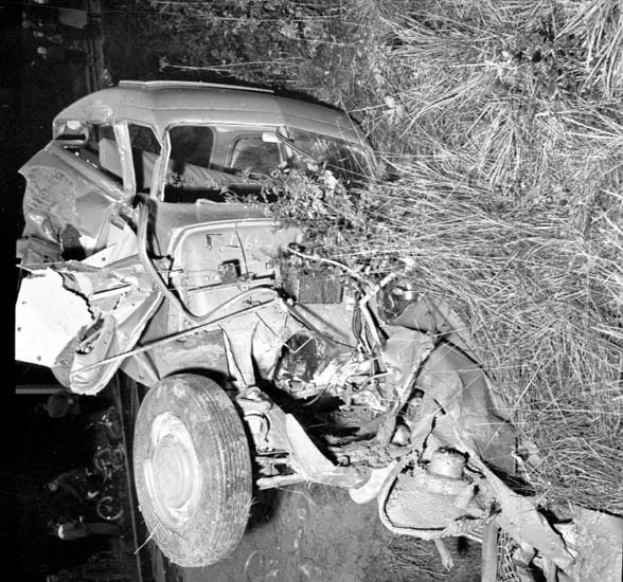 Ein Schwarzweißfoto eines schwer beschädigten Autos am Straßenrand, mit einer eingedrückten Front, zersplitterten Fenstern, einem Baum im Hintergrund und verstreuten Gegenständen an der Szene.