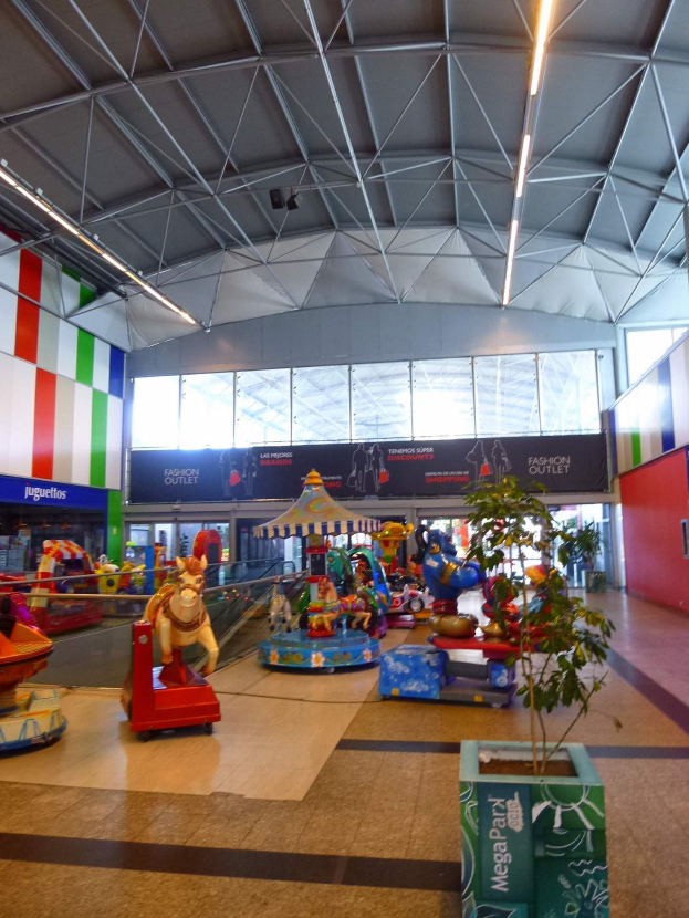 Vergnügungsgeräte, eine Topfpflanze, verstreute Spielzeuge, Informationsschilder, eine Wand, Deckenleuchten und Metallstangen in einem großen Indoor-Einkaufszentrum.