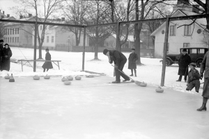Schwarzes Bild von Menschen, die Curling auf einem Outdoor-Eisstadion spielen, das von einem Zaun umgeben ist, mit einer Bank, Bäumen, Gebäuden und einem geparkten Fahrzeug im Hintergrund.