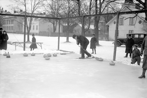 Schwarzes Bild von Menschen, die Curling auf einem Outdoor-Eisstadion spielen, das von einem Zaun umgeben ist, mit einer Bank, Bäumen, Gebäuden und einem geparkten Fahrzeug im Hintergrund.