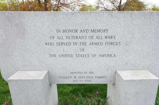 Ein Gedenkstein mit einer Inschrift, die allen US-Veteranen gewidmet ist, umgeben von Pflanzen, Blumen, Gras, Bäumen und unter einem bewölkten Himmel.