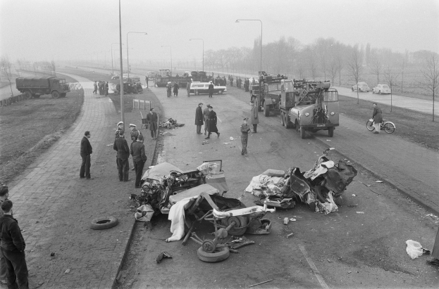 Schwarze und weiße Szene eines Autounfalls am Straßenrand mit mehreren Fahrzeugen und einer Gruppe von Menschen drumherum, Laternen, Bäume und Himmel im Hintergrund.