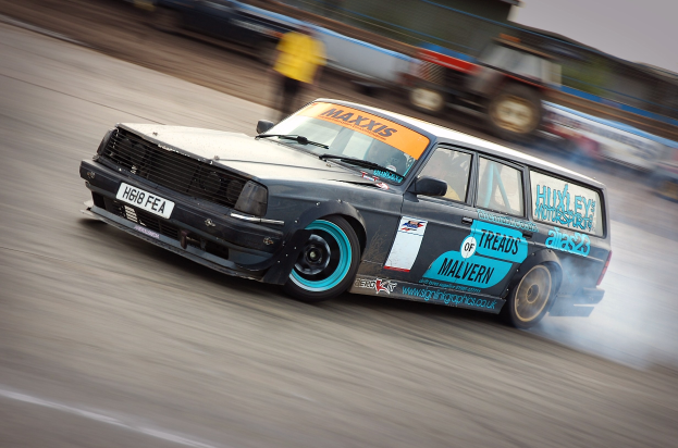 Ein Auto rast eine Rennstrecke entlang und hinterlässt eine Rauchspur, umgeben von anderen Fahrzeugen und Menschen im Hintergrund.