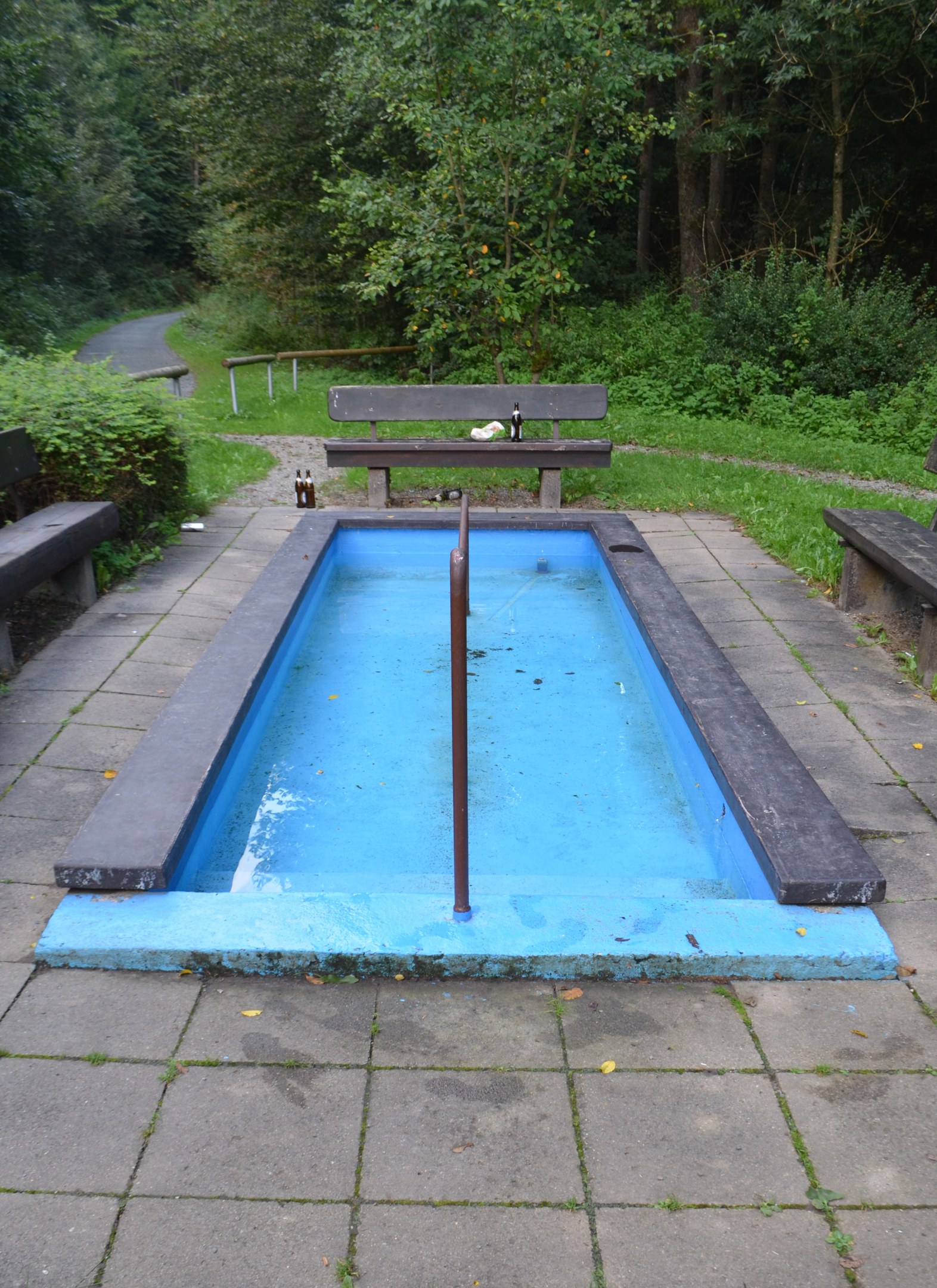 Kleiner Parkpool mit zentralem Pfahl, umgeben von Bänken, Grün, Straße und Bäumen.