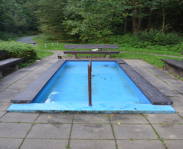 Kleiner Parkpool mit zentralem Pfahl, umgeben von Bänken, Grün, Straße und Bäumen.