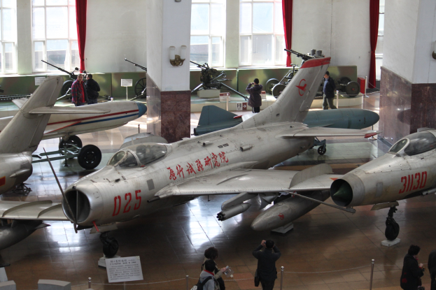 Eine Gruppe von Kampfflugzeugen in einem Museum ausgestellt, mit Besuchern, die hinter Barrieren und Informationsschildern beobachten.