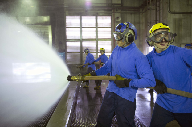 Gruppe von Männern in blauen Hemden und gelben Helmen, die an einer Maschine arbeiten, wobei einer einen Schlauch hält und Wasser auf den Boden sprüht, in einer Fabrik mit sichtbaren Rohren, Fenstern und Lampen.