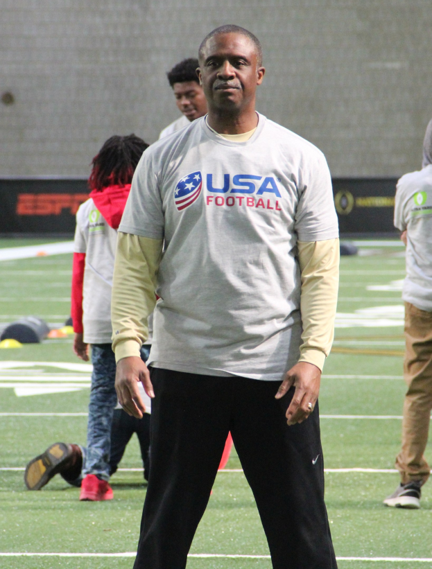 Ein Fußballspieler in Fußballschuhen steht auf einem Feld mit einer Gruppe von Menschen und einer Wand im Hintergrund.