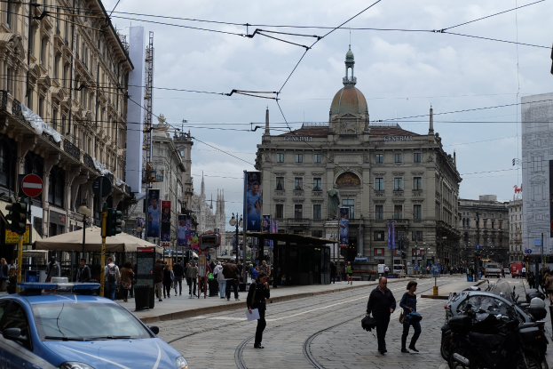 Eine belebte Stadtstraße mit einem parkenden Polizeiwagen, Fußgänger mit Taschen, fahrende Fahrzeuge, hohe Gebäude mit Fenstern, Banner, Laternenpfähle, Ampeln und ein bewölkter Himmel.