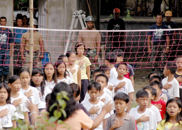 Kinder vor einem Netz stehend, Frau im Vordergrund, Menschen mit Helmen, Leiter, Wand und Bäume im Hintergrund