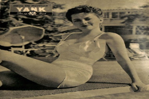 Olivia de Havilland in einem Badeanzug auf dem Boden sitzend und lachend, mit Bäumen, Stühlen und einem Gebäude im Hintergrund und Text oben.
