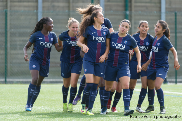 Eine Gruppe junger Frauen, die auf einem Rasenfeld Fußball spielen, mit Maschendrahtzaun und einer Wand im Hintergrund, mit dem Text "Paris Saint-Germain Frauenfußball" in der rechten unteren Ecke.