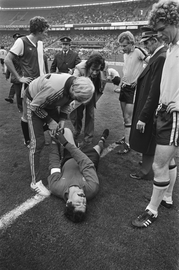 Schwarzes und weißes Bild eines Fußballspielers, der auf dem Boden liegt und von einer Gruppe Männer umgeben ist, während eine Stadionmenge zusieht.