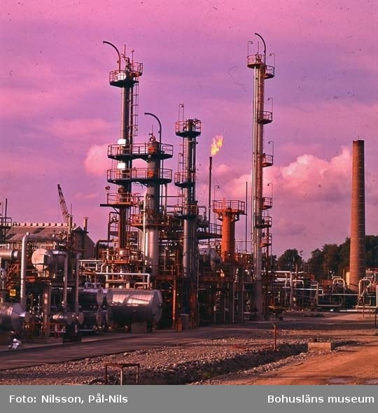 Industrielle Ölraffinerie mit ausgedehnten Rohrleitungen, Metallstrukturen, Containern, Bäumen, einem Gebäude, einem Kran und einem bewölkten Himmel mit Text unten.