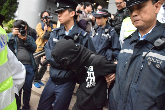 Eine Gruppe von Polizeibeamten steht vor einer Menge, einige halten Kameras, mit einem Gebäude mit Säulen und Bäumen im Hintergrund.