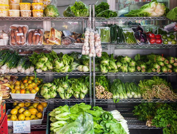 Ein Gang in einem Supermarkt mit frischem Gemüse und Obst in Plastikverpackungen, Kartons, einer Texttafel und einem Glasfenster im Hintergrund.