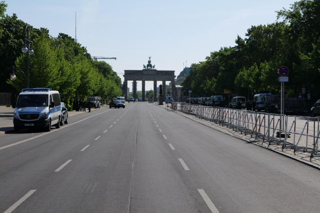 Ein Polizeiwagen steht auf der Seite einer vielbefahrenen Straße vor dem Brandenburger Tor in Berlin, Deutschland, mit Bäumen, Laternenmasten, Absperrungen und Schildern im Hintergrund und einer bewölkten Himmel.