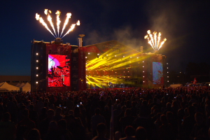 Eine große Menschenmenge auf einem Musikfestival mit einer Bühne in der Mitte, Musiker unter hellen Scheinwerfern, Banner mit Text, Zelte links und ein dunkler Himmel, der von Feuerwerk erhellt wird.