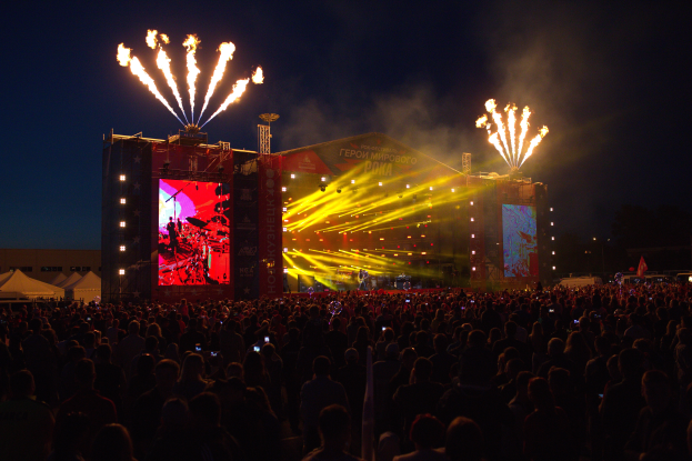 Eine große Menschenmenge auf einem Musikfestival mit einer Bühne in der Mitte, Musiker unter hellen Scheinwerfern, Banner mit Text, Zelte links und ein dunkler Himmel, der von Feuerwerk erhellt wird.