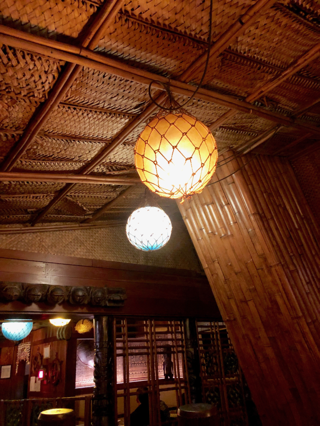 Ein Restaurant-Innenraum mit einer hölzernen Decke, an der Lampen hängen, Tischen und Stühlen, einer Person, die sitzt, und Wänden, die mit Foto-Rahmen und anderen Gegenständen dekoriert sind.