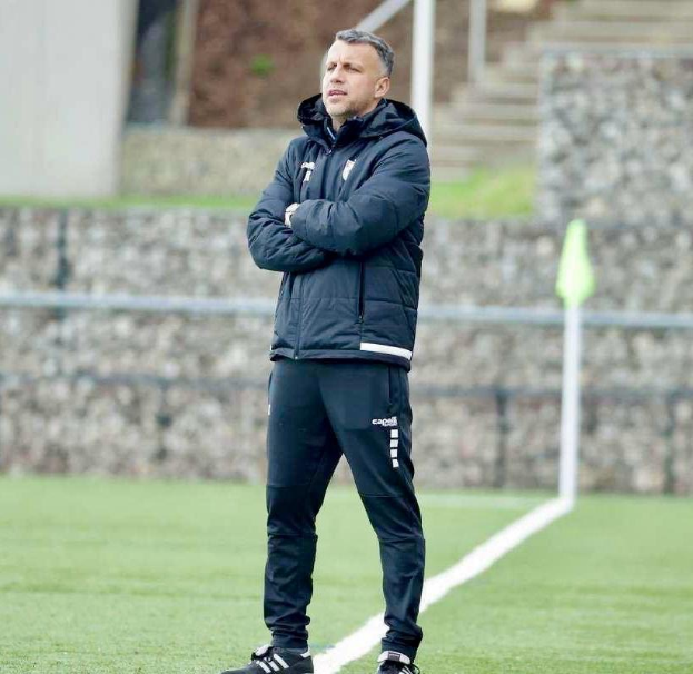 Ein Fußballtrainer mit verschränkten Armen auf einem Feld stehend, der eine schwarze Jacke und Hose trägt, mit einem Zaun, Treppe und Wand im Hintergrund.