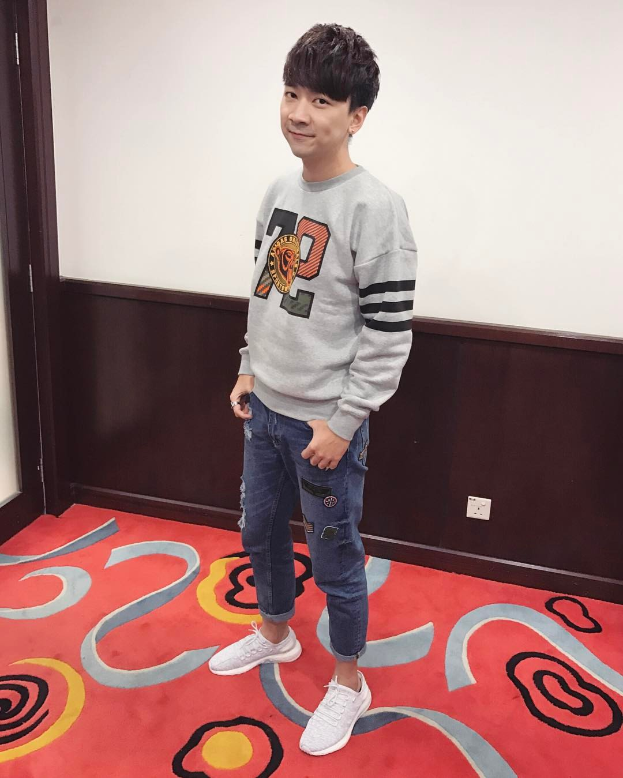 Ein junger Mann steht auf einem roten Teppich bei einer koreanischen Modenschau, trägt ein graues Sweatshirt und blaue Jeans und lächelt mit einer Schalttafel im Hintergrund.