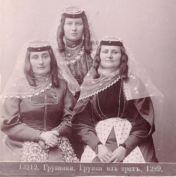 Schwarzes und weißes Foto von drei Frauen in traditionellen russischen Hochzeitskleidern mit farbenfrohen Stickereien und Perlenoutfits, die vor einer weißen Wand stehen.