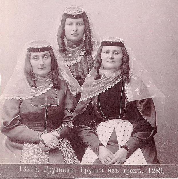 Schwarzes und weißes Foto von drei Frauen in traditionellen russischen Hochzeitskleidern mit farbenfrohen Stickereien und Perlenoutfits, die vor einer weißen Wand stehen.