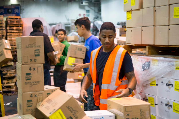 Eine Gruppe von Männern in orangefarbenen Westen und Sicherheitswesten lädt Kisten in ein Lagerhaus, mit mehreren Kartonboxen rechts und einer Kette links und einer Wand mit Lampen im Hintergrund.