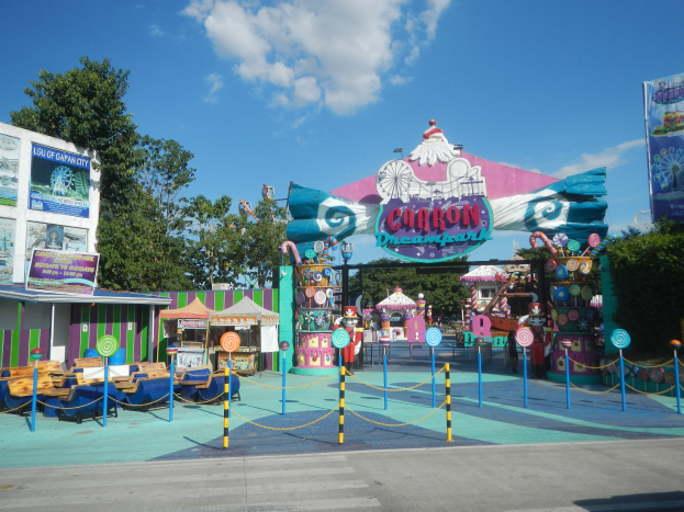 Karussell am Parkeingang mit Straße, eingezäuntem Bereich, Buden, Schildern, Bäumen und bewölktem Himmel.
