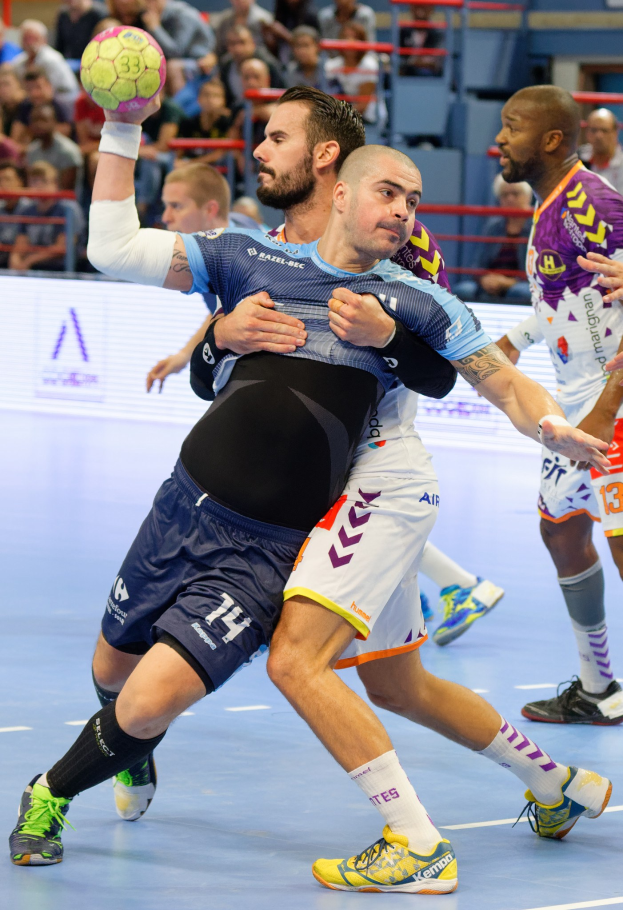 Mannergruppe beim Handballspielen in einem Stadion, einer hält den Ball, mit Zuschauern im Hintergrund.