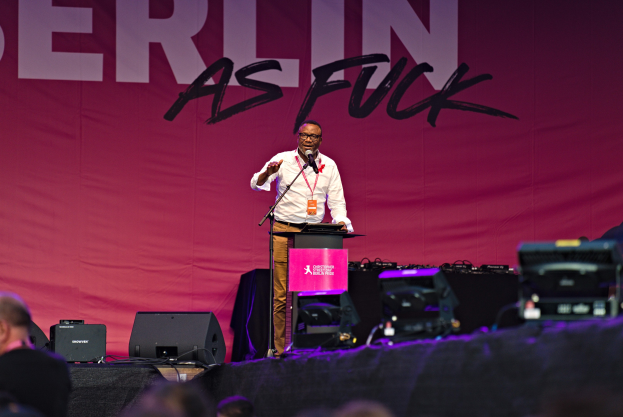 Mann in weißem Hemd und braunen Hosen, der in ein Mikrofon spricht, vor einem pinken 'Berlin as Fuck'-Plakat auf der Bühne steht.