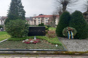Ein Denkmal für die Opfer des Zweiten Weltkriegs steht in einem Park, geschmückt mit einem Kränzchen und umgeben von Grün, mit Gebäuden im Hintergrund.