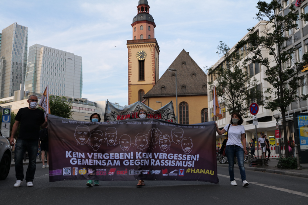 Gruppe von maskierten Menschen auf der Straße mit einem Banner, daneben ein geparktes Auto, im Hintergrund Gebäude und ein Uhrenturm unter einem klaren blauen Himmel.
